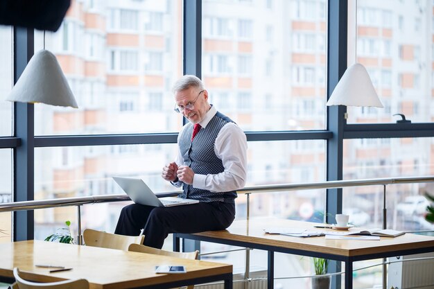Uomo d'affari maschio adulto, insegnante, mentore che lavora a un nuovo progetto. si siede vicino a una grande finestra sul tavolo. guarda lo schermo del laptop.