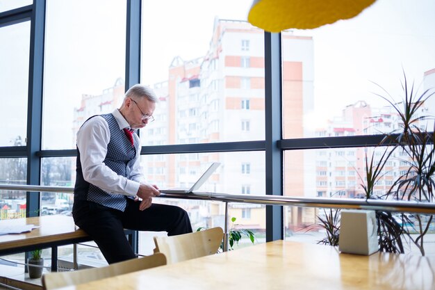 Adult male businessman, teacher, mentor working on a new project. Sits by a large window on the table. He looks at the laptop screen.