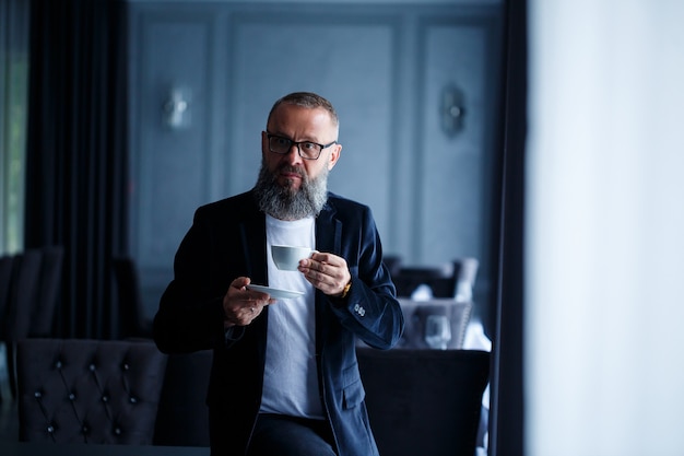 An adult male mentor, director, businessman in glasses and a suit drinks coffee and has a rest. Working day concept