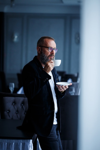 An adult male mentor, director, businessman in glasses and a suit drinks coffee and has a rest. Working day concept