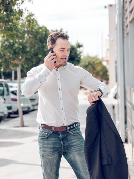 Adult male worker in white shirt and jeans checking time and talking on smartphone before working meeting