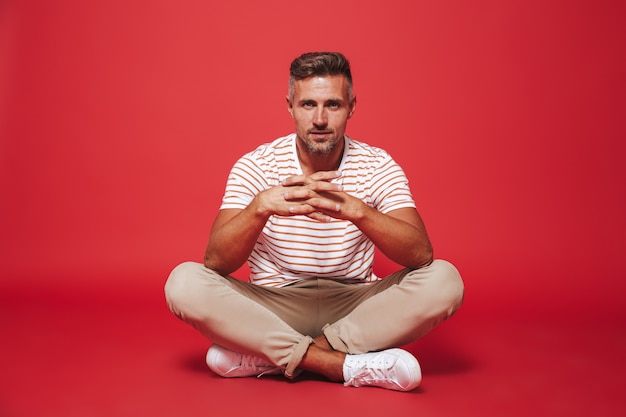 adult man 30s in striped t-shirt smiling, while sitting on floor with legs crossed isolated on red