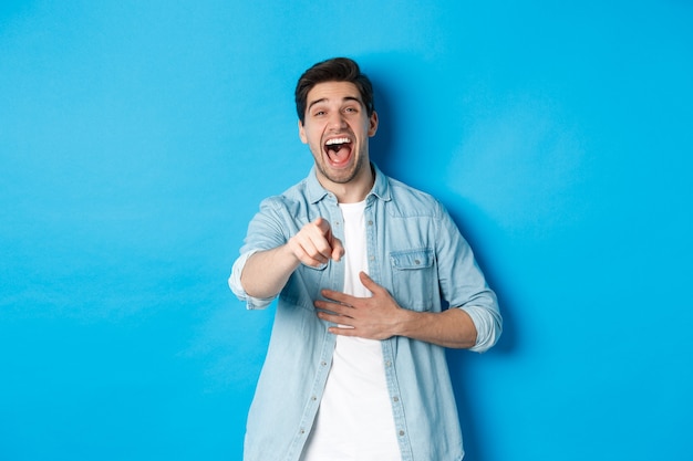 Adult man in casual outfit laughing out loud and pointing at you, looking at something funny, chuckling while standing against blue background