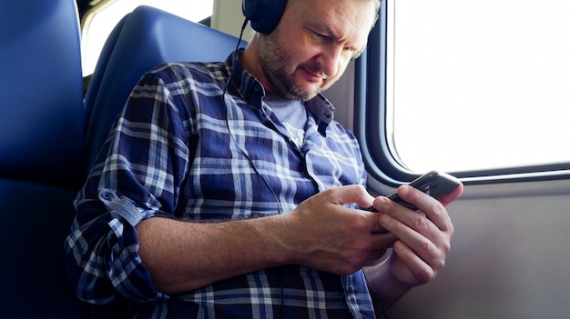 An adult man in a commuter train with headphones looks at the phone and listens to music