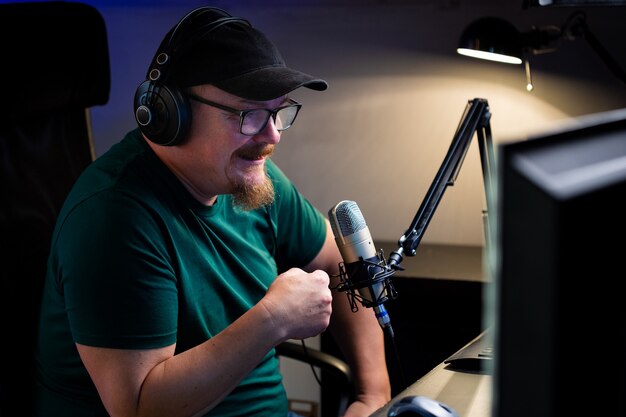 Фото Взрослый мужчина в очках с бородаем ведет прямую трансляцию через интернет. живой блоггер разговаривает с камерой на компьютере с мониторами. говорит в микрофон.