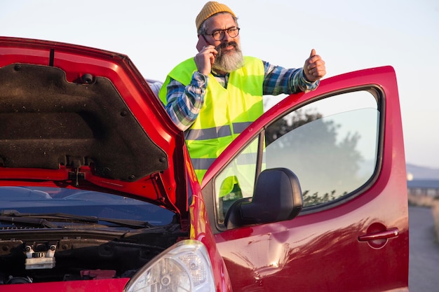 Adult man looking calm and talking on the phone on the side of\
the road. latino man with hood open due to mechanical problem