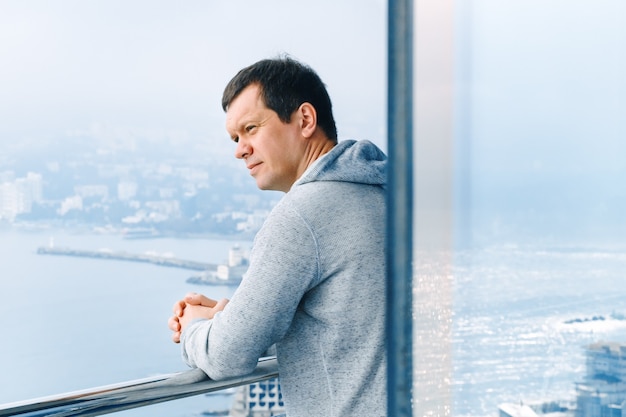 An adult man looks at the sea and stands on the balcony of a glass building