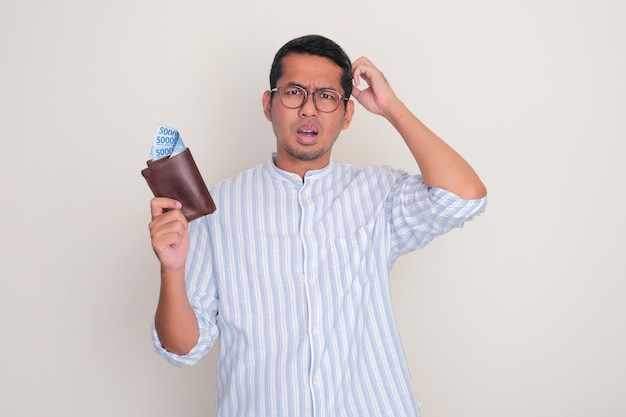 Adult man scratching head showing confused expression while holding wallet full of money