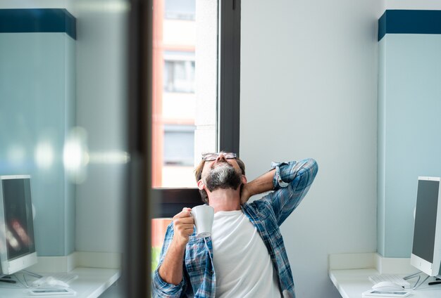 仕事の姿勢によって引き起こされるストレスの痛みや病気のために首と背中に触れる成人男性-成人男性は、職場の座席に座って背中の問題に苦しんでいます-フリーランスの疲れた人々のライフスタイル