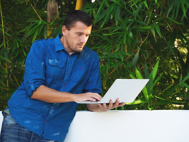 Adult man uses a laptop against green leaves.