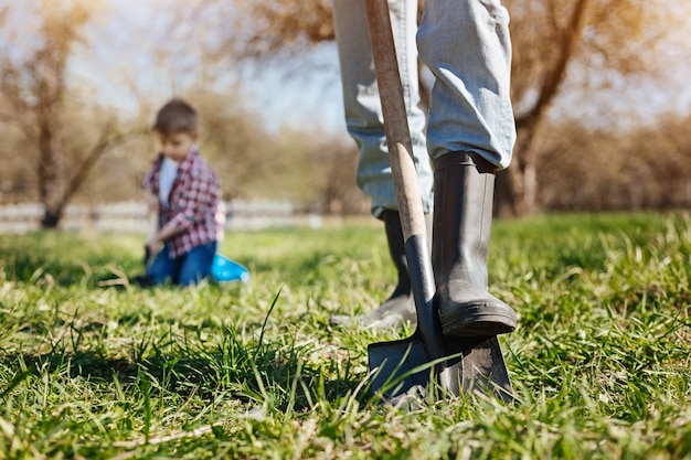 Adult man wearing green wellies digging the soil with a shovel while spending free time outside with his grandson