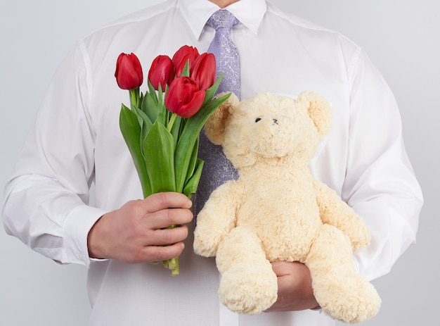 L'uomo adulto in una camicia bianca tiene un mazzo dei tulipani di fioritura rossi e un orsacchiotto bianco