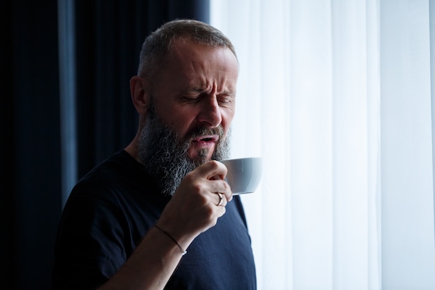 An adult man with a beard drinks coffee and looks out the window. Working day field recreation concept