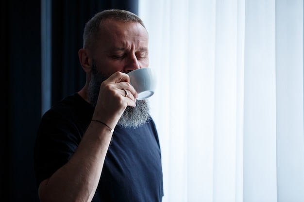 An adult man with a beard drinks coffee and looks out the window. Working day field recreation concept