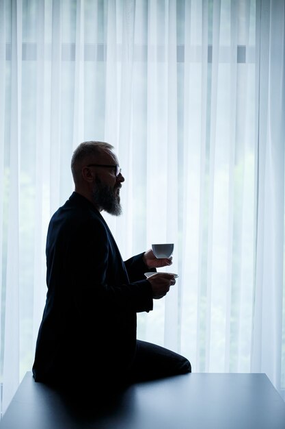 An adult man with a beard drinks coffee and looks out the window. Working day field recreation concept