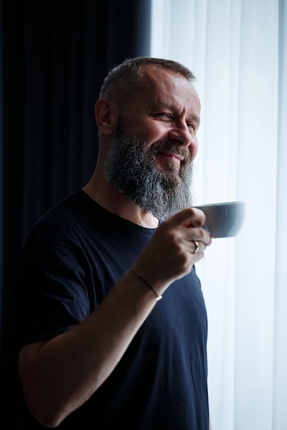An adult man with a beard drinks coffee and looks out the window. Working day field recreation concept