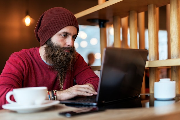 Adult man with braided beard sitting in cafe having cup of coffee while typing on laptop