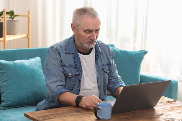 Adult man with gray hair sitting on sofa at home, and working on laptop