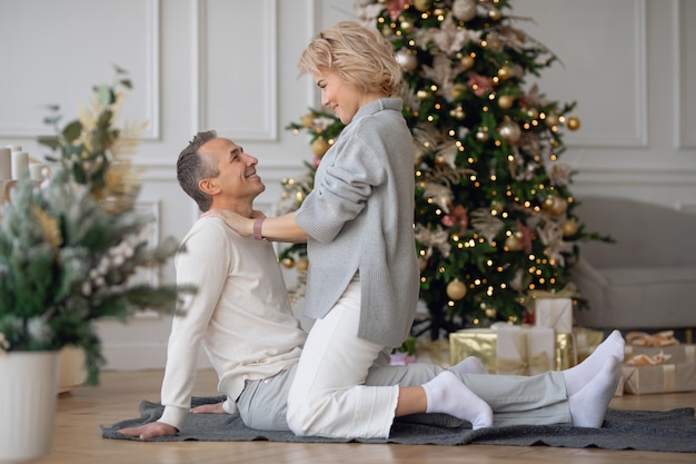 adult man and woman sit on the floor near the christmas tree and hug