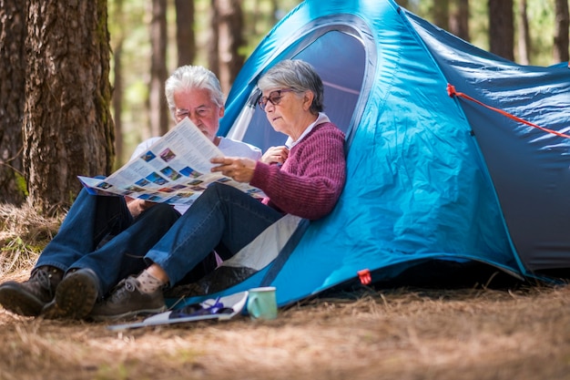 Adult mature retired couple enjouying the wild camping outdoor in the forest looking together a paper map to choose the next adventure destination to see and to live - travel tourism concept