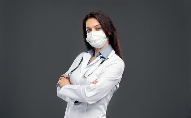흰 가운과 수술용 마스크를 쓴 성인 의료 종사자는 팔짱을 끼고 회색 배경에 대한 코로나바이러스 전염병 동안 카메라를 보고 있습니다.