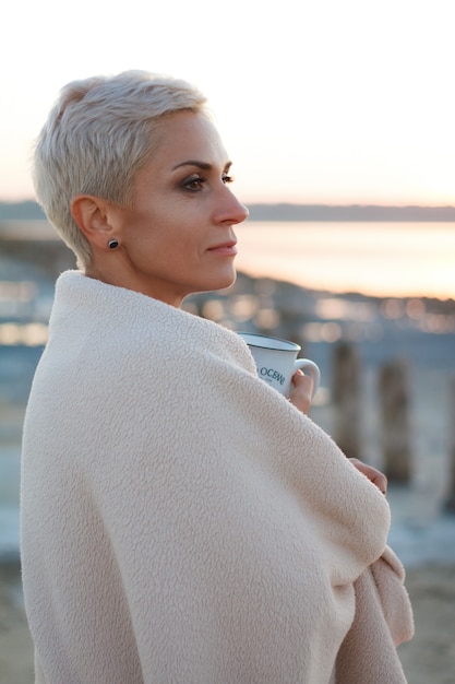 Adult middle-aged blonde woman wrapped herself in a bright plaid on the beach and looks at the sea