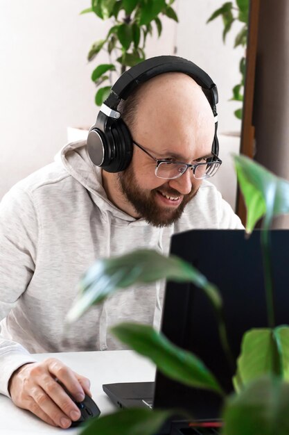 Adult millenial man with glasses and headphones sitting at table working online on laptop at home