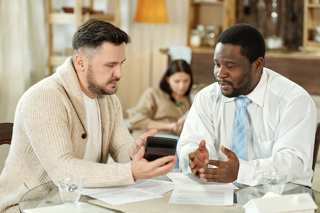 Adult multiethnic man and financial consultant sitting at table with calculating machine while discussing mortgage