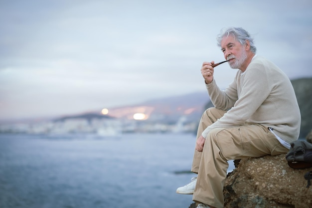 Adult old senior bearded man resting during excursion at sea smoking pipe looking at horizon over water Caucasian elderly whitehaired male enjoying free time vacation and nature in seascape