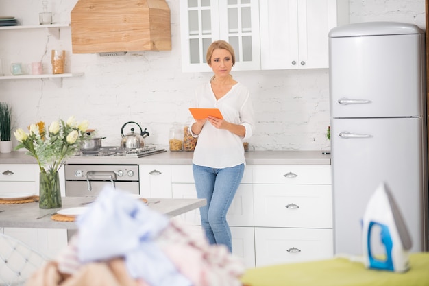 Adult pretty woman in jeans with tablet in her hands standing at home in kitchen