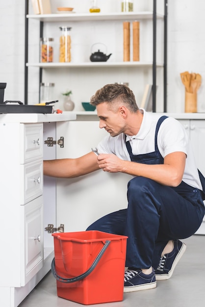 Adult repairman with red plastic bucket and spanner repairing kitchen sink