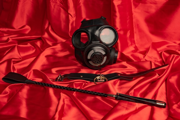 Adult sex games. BDSM items. Gas mask, collar and whip on a red satin sheet.