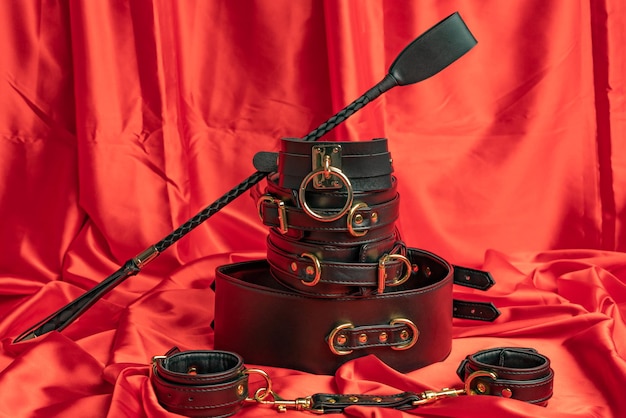 Adult sex games. BDSM items. Leather straps handcuffs, collar, belt and whip on a red satin sheet.