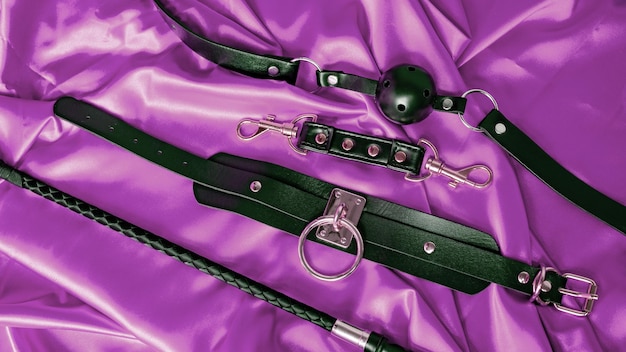 Adult sex games BDSM items Leather straps handcuffs collar gag ball and whip on a purple satin sheet