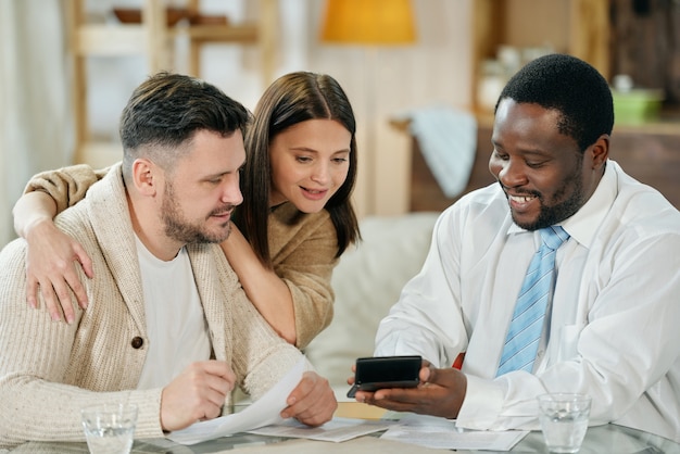 Uomo di colore sorridente adulto con la macchina calcolatrice che mostra alle giovani coppie il loro tasso di interesse