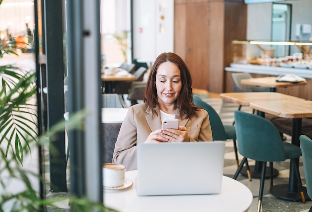 Adult smiling brunette business woman in beige suit working on laptop drinking coffee at cafe