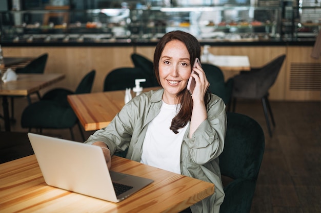 Adult smiling brunette business woman forty years with long hair in stylish shirt working on laptop using mobile phone in cafe