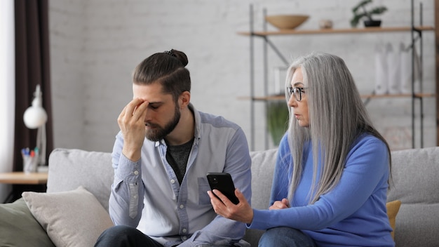 Photo adult son teaching his senior mother to use smartphone mobile apps sharing photos shopping online