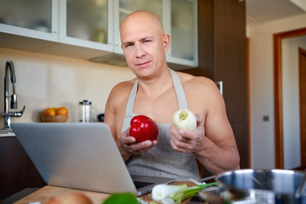 Adult strong man in kitchen preparing food and looks into laptop