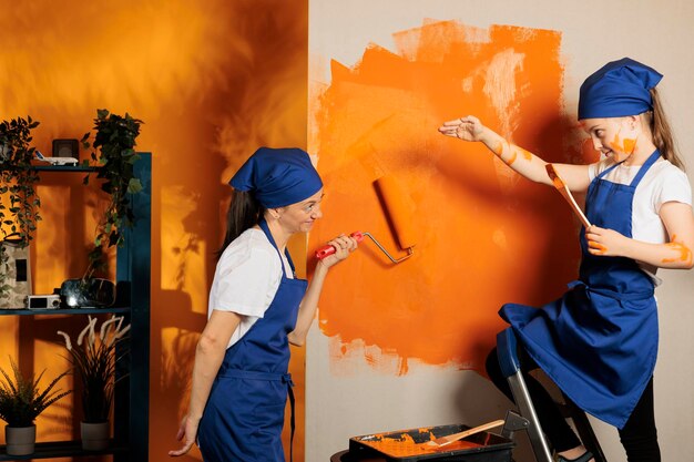 Adult with kid painting wall with orange paint, using\
renovating diy tools to change surface color in house apartment.\
small child using paintbrush on ladder and mother holding roller\
brush.