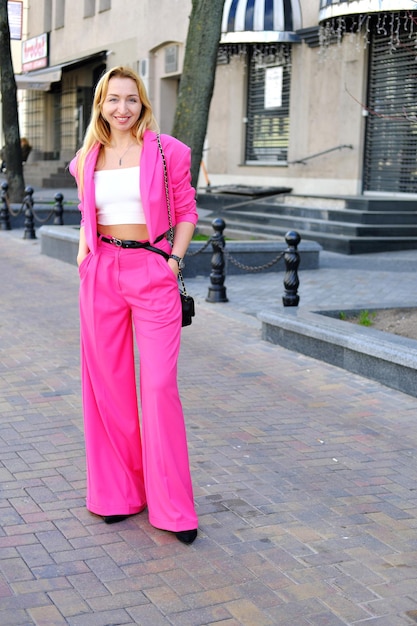 An adult woman in a bright pink trouser suit and a white top poses on a city street