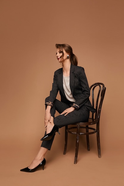 Adult woman caucasian business lady in a grey suit blouse and black shoes posing on the chair over beige wall isolated with copy space