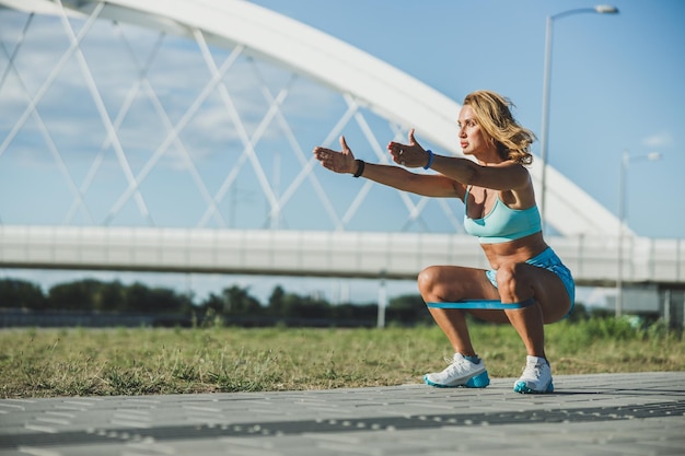 Adult woman doing squats with resistance band on her legs while working out near the river.