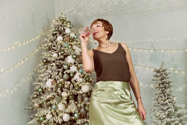 An adult woman drinks champagne next to a christmas tree on new years eve
