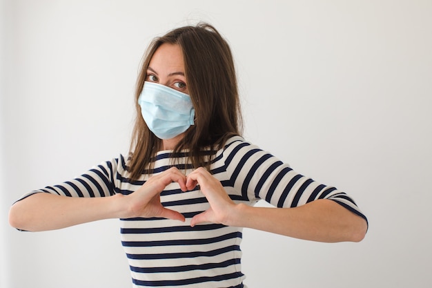 Взрослая женщина в маске смотрит в камеру и жестикулирует сердце возле груди во время пандемии против серого