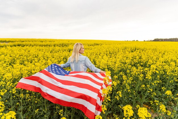 Adult woman holding American flag with pole, stars and stripe in a yellow rapeseed field. USA flag fluttering in the wind.