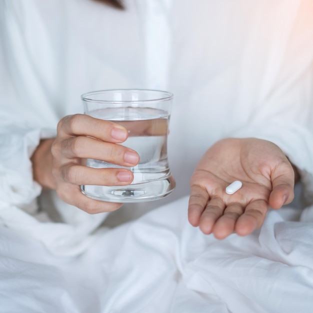 Adult woman holding pill and glass of water female taking medicine on bed at home Migraine painkiller headache influenza illness sickness and healthcare concept