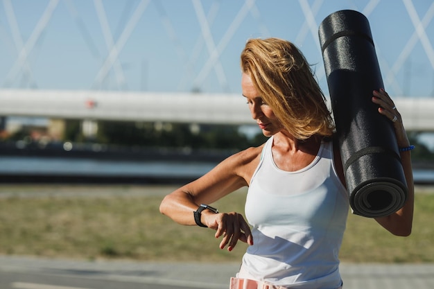 Adult woman is resting after hard training near the river and looking on smartwatch.