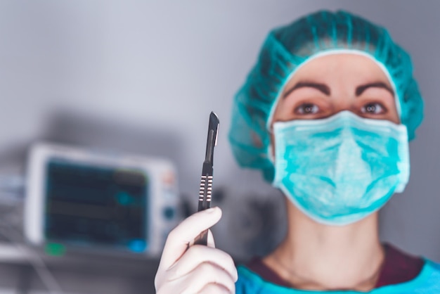 Взрослая женщина в медицинской маске и шляпе и со скальпелем смотрит в камеру перед операцией в больнице Фото высокого качества
