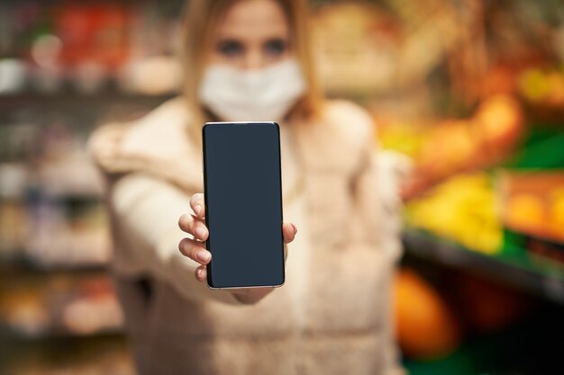 Foto donna adulta in maschera medica che utilizza smartphone e fa la spesa